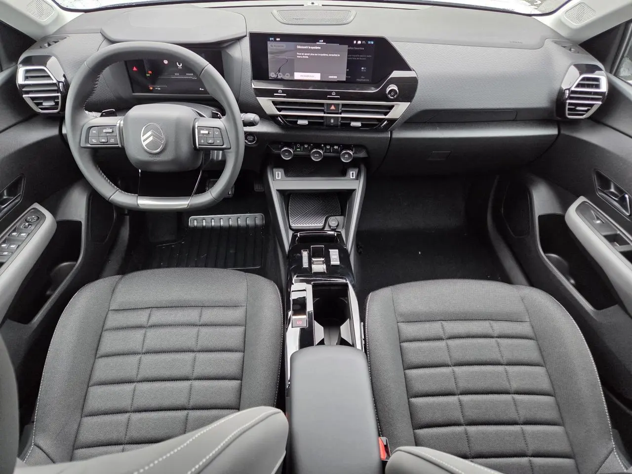 Vue intérieure avant de la Citroën C4 2025, tableau de bord moderne avec écran tactile et sièges tissu noir.