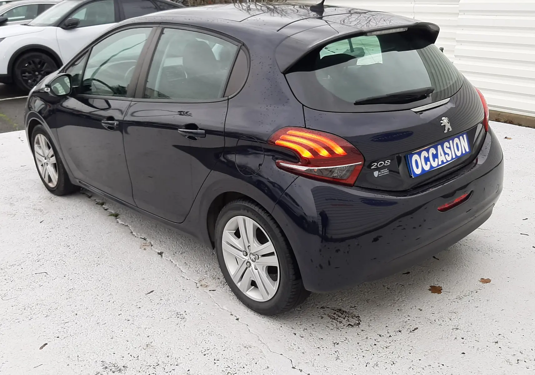 Peugeot 208 bleu nuit vue 3/4 arrière droit, feux arrière allumés et plaque d'occasion visible.