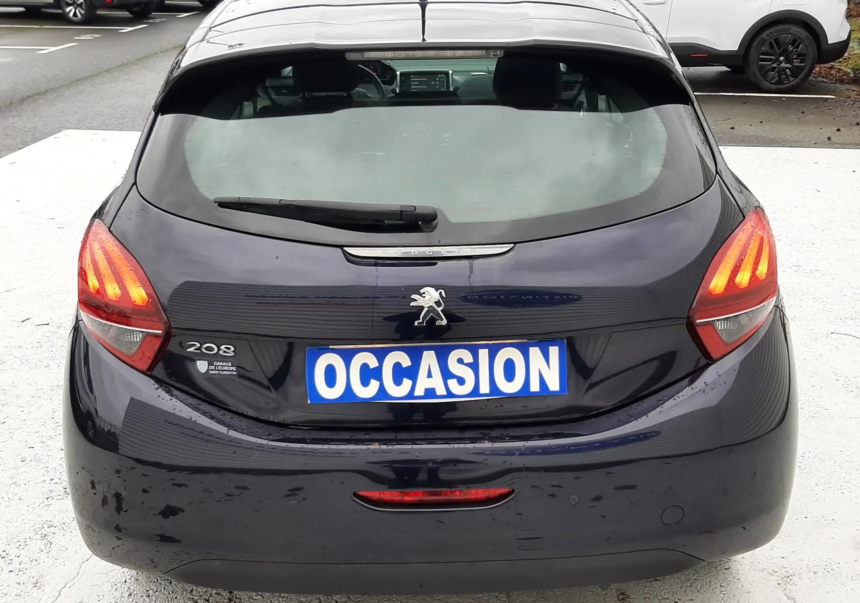 Vue arrière d'une Peugeot 208 bleu nuit 2019 avec feux allumés et plaque "OCCASION" visible.