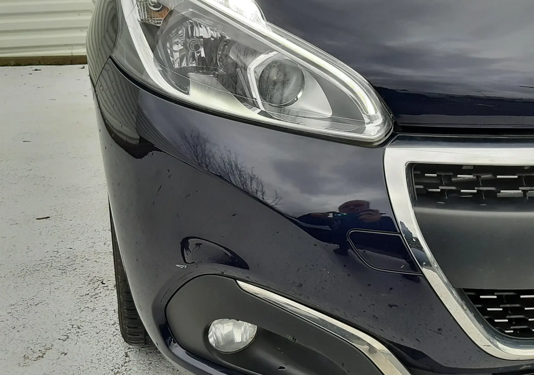 Gros plan sur l'avant gauche d'une Peugeot 208 bleu nuit 2019, mettant en valeur le phare LED et la calandre chromée.