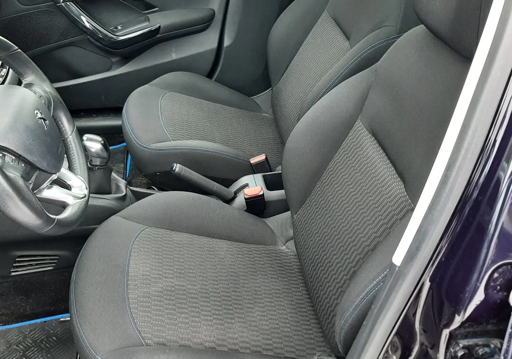 Vue intérieure côté conducteur de la Peugeot 208 bleu nuit 2019, sièges tissu noir avec surpiqûres bleues et volant compact.