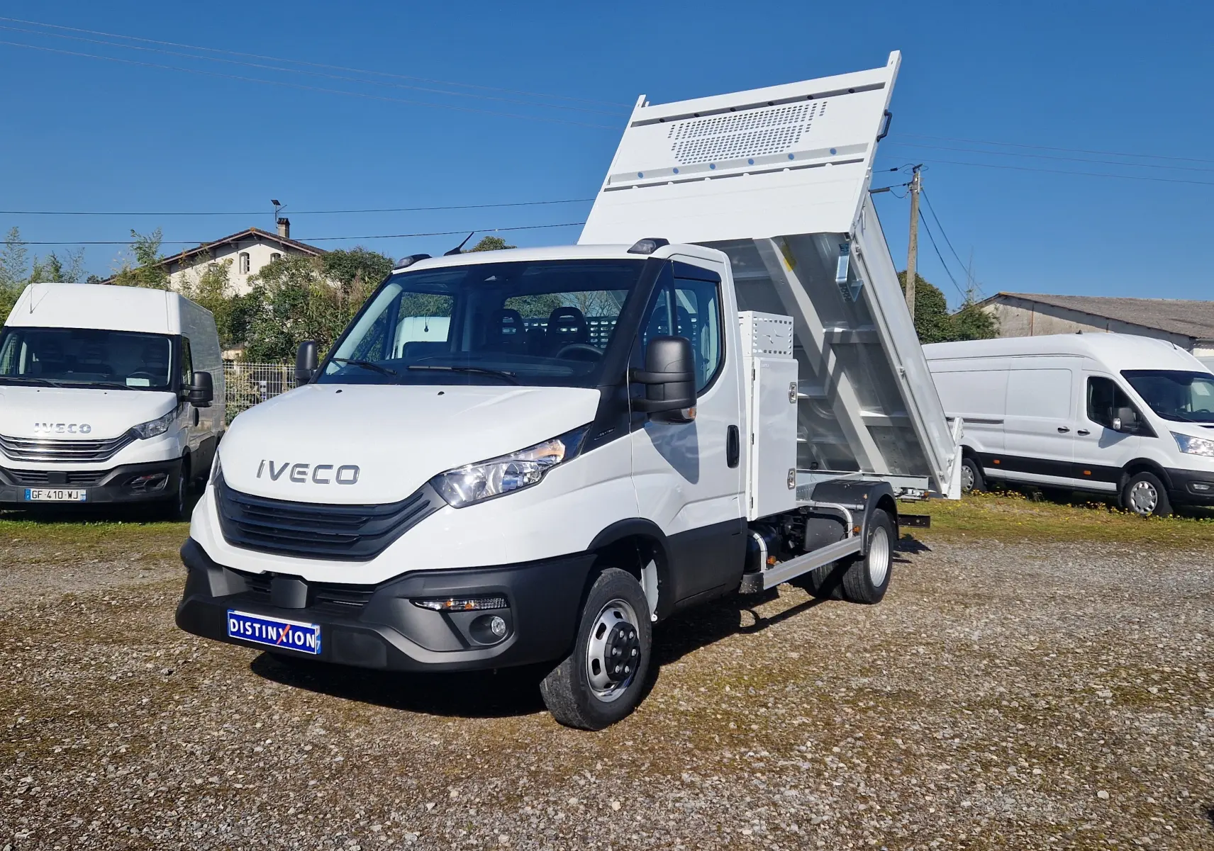IVECO Daily blanc en 3/4 avant droit avec benne relevée et coffre latéral visible sur terrain extérieur.