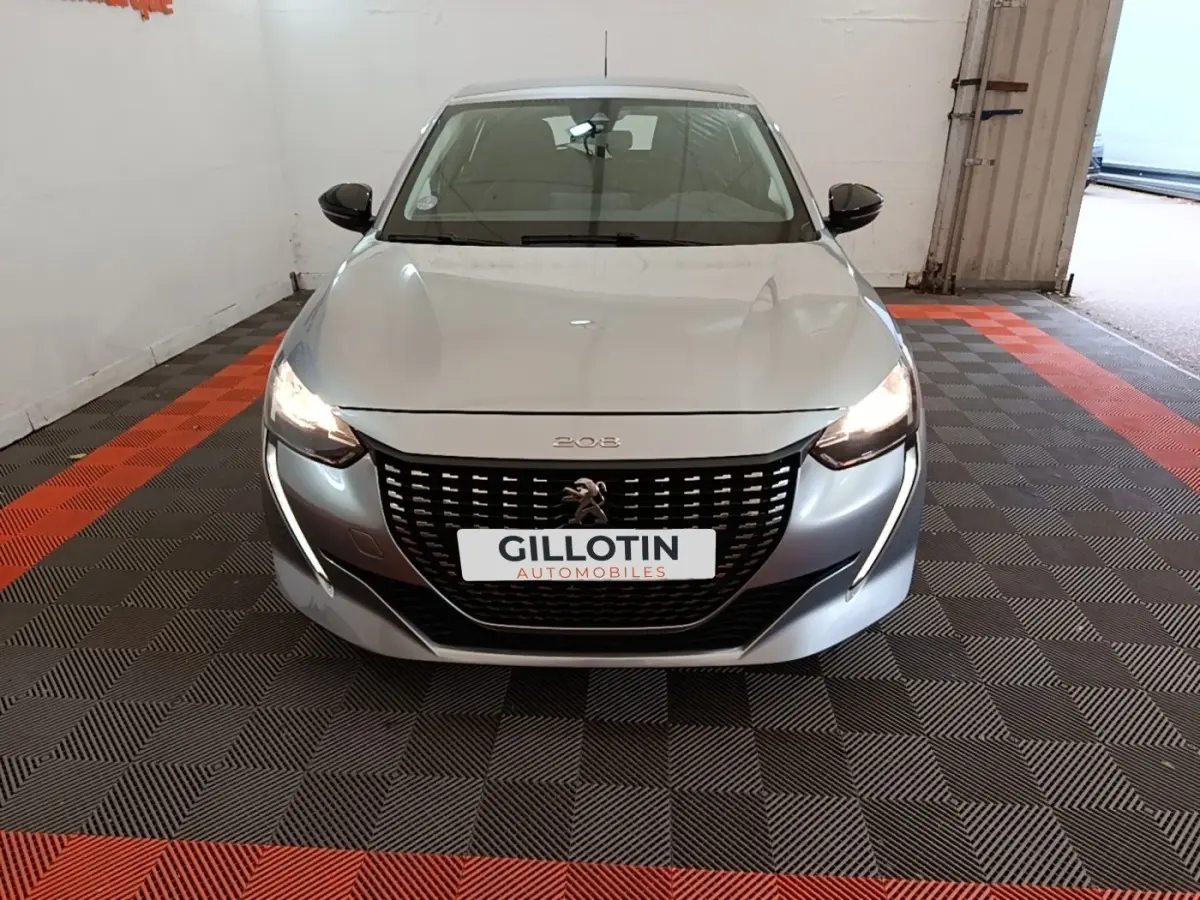 Vue avant d'une Peugeot 208 gris clair avec phares allumés dans un garage fermé.