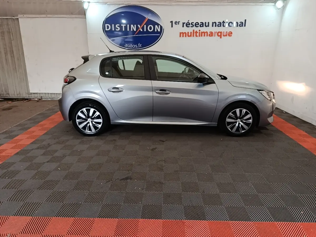 Profil droit d'une Peugeot 208 gris clair 2023 garée en intérieur sur sol à damier noir et rouge.