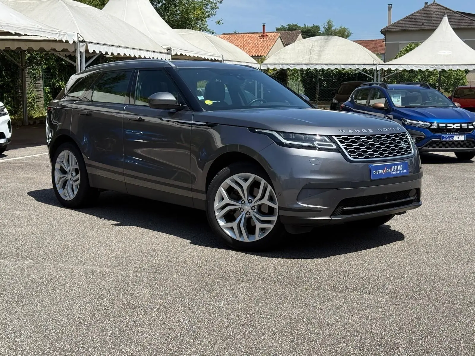 Range Rover Velar gris en 3/4 avant droit, avec jantes alliage et calandre distinctive sur parking extérieur.