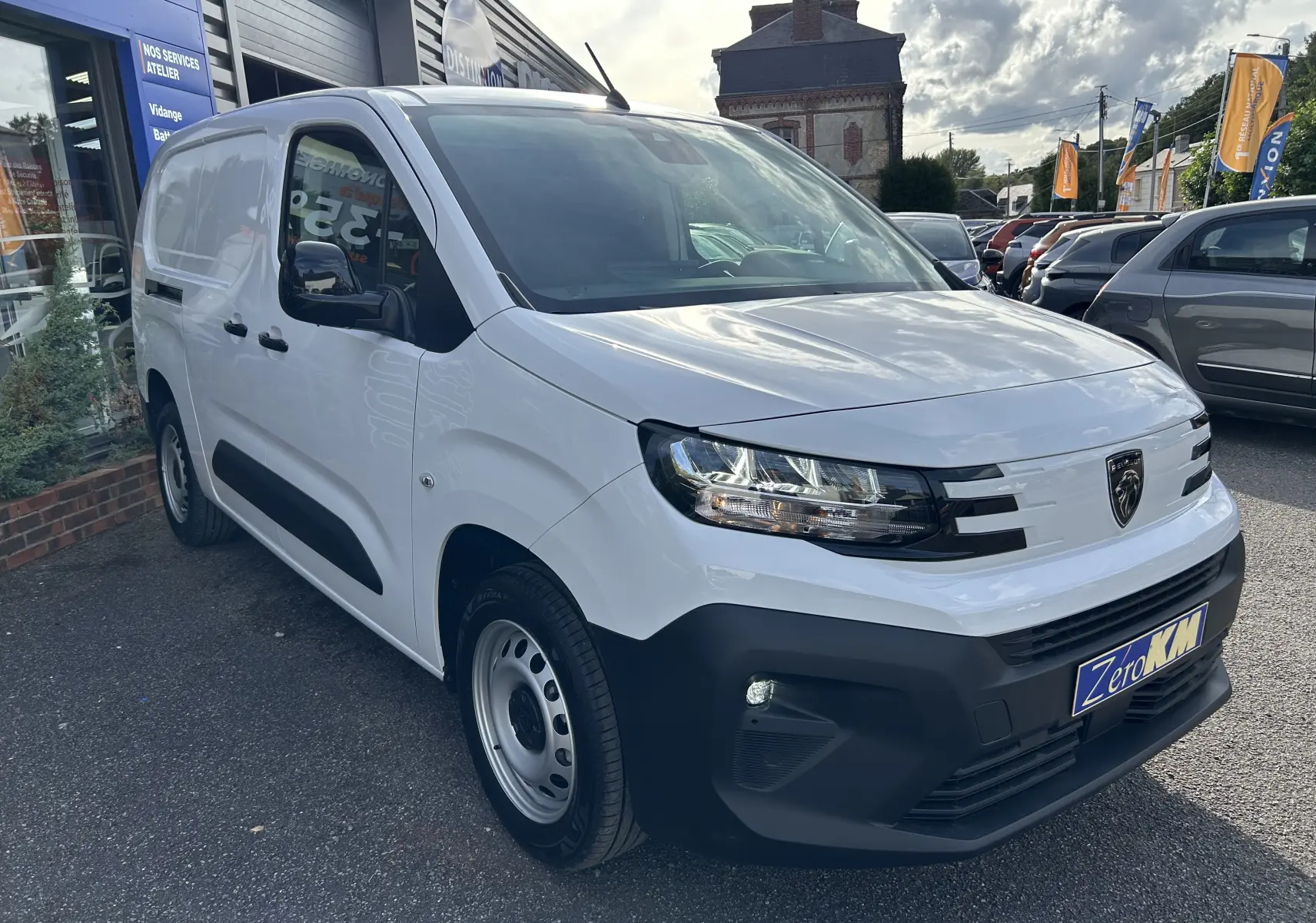 Peugeot Partner Fourgon blanc en 3/4 avant droit avec rétroviseurs noirs et calandre moderne, stationné devant un atelier.