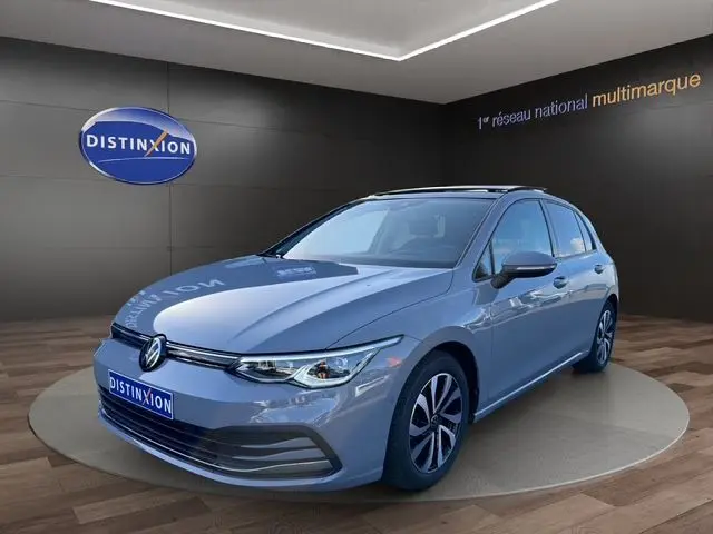 Volkswagen Golf VIII gris lunaire vue 3/4 avant en intérieur avec jantes alu et toit panoramique visible.