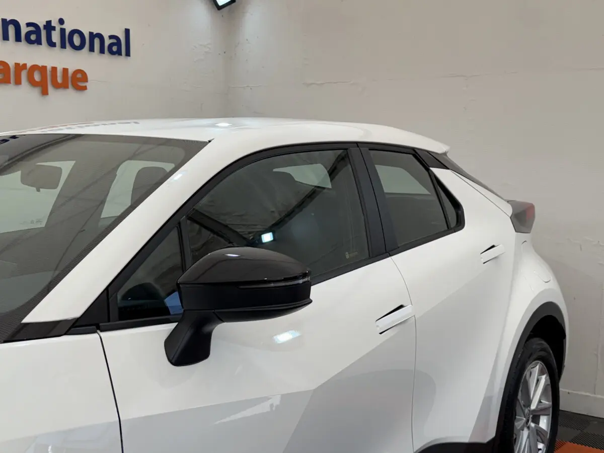 Vue de profil côté gauche d'un Toyota C-HR 1.8 Hybride blanc avec rétroviseur noir en intérieur showroom.