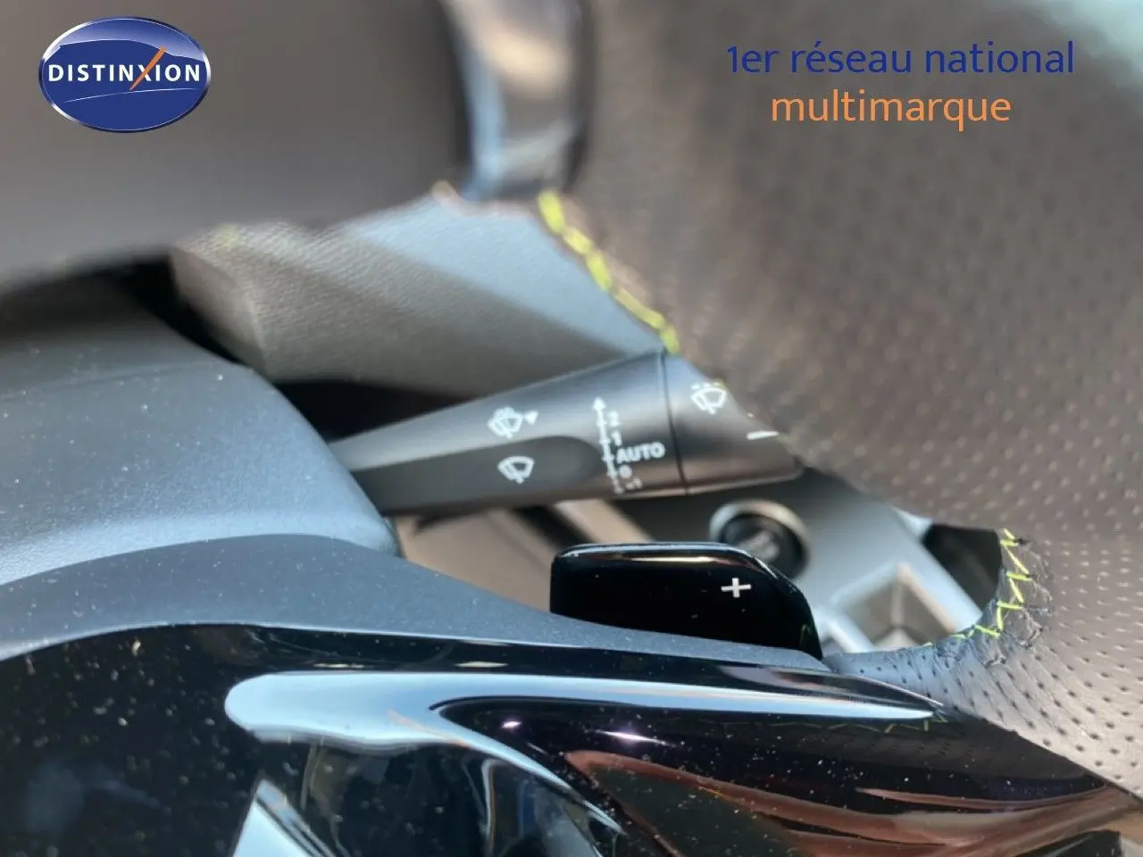 Gros plan sur la palette de changement de vitesse droite en plastique noir brillant, volant cuir perforé avec surpiqûres jaunes.
