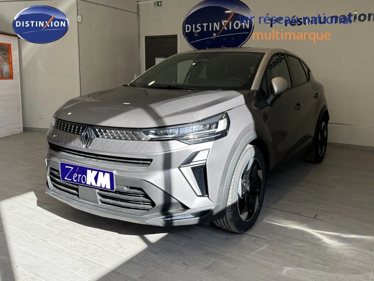 Vue 3/4 avant du Renault Captur 2025 gris Cassiopée métal avec toit gris, en intérieur showroom, mettant en valeur ses optiques LED et jantes biton.