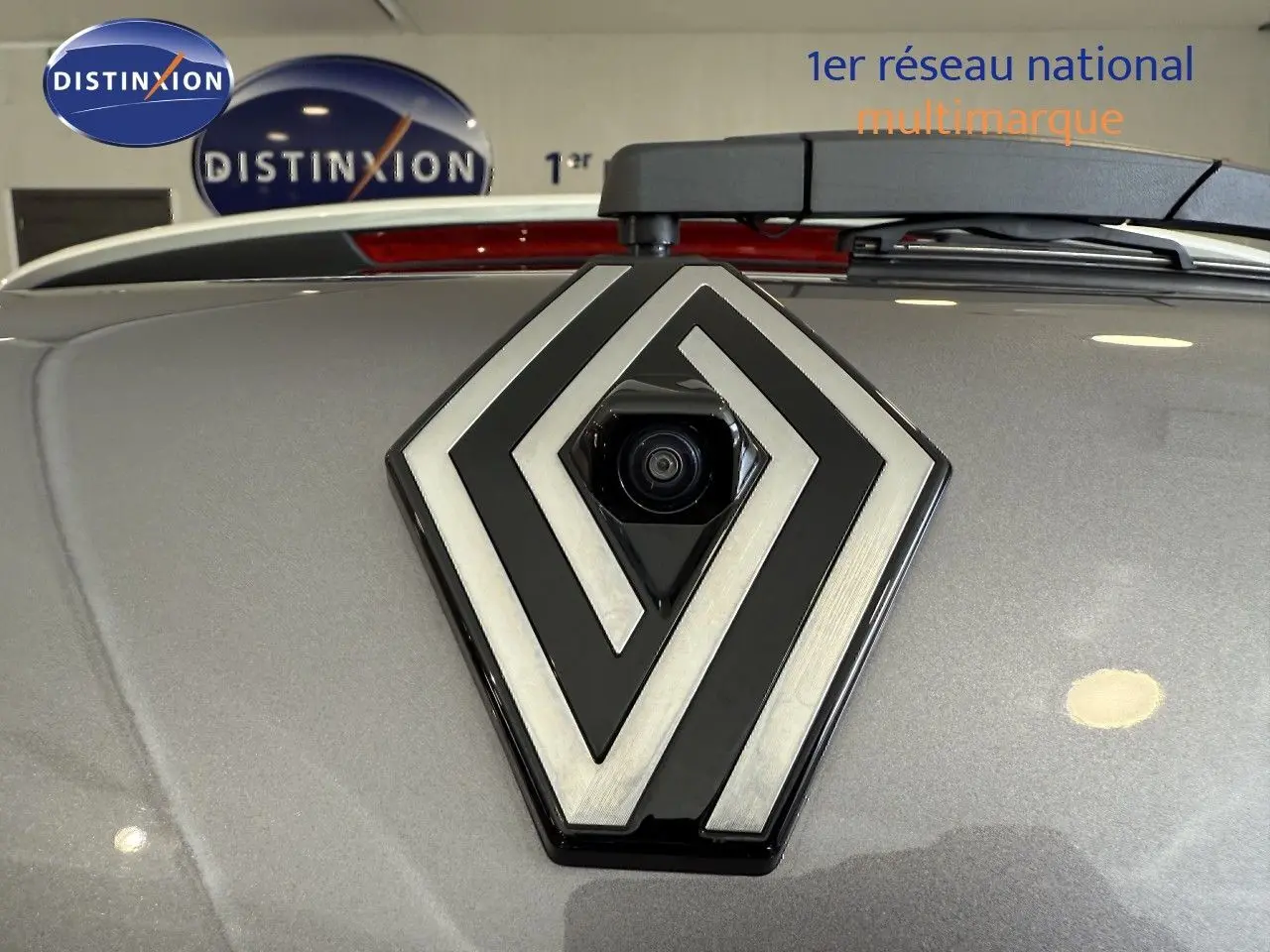 Gros plan sur la caméra arrière intégrée dans le logo Renault sur un capot gris cassiopée métallisé.
