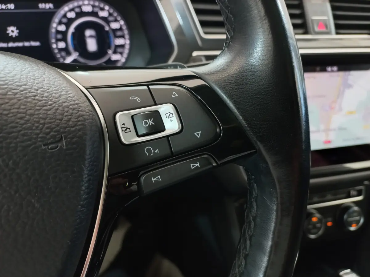 Gros plan sur les commandes multifonctions côté gauche du volant cuir noir du Volkswagen Tiguan Allspace marron, avec tableau de bord numérique en arrière-plan.