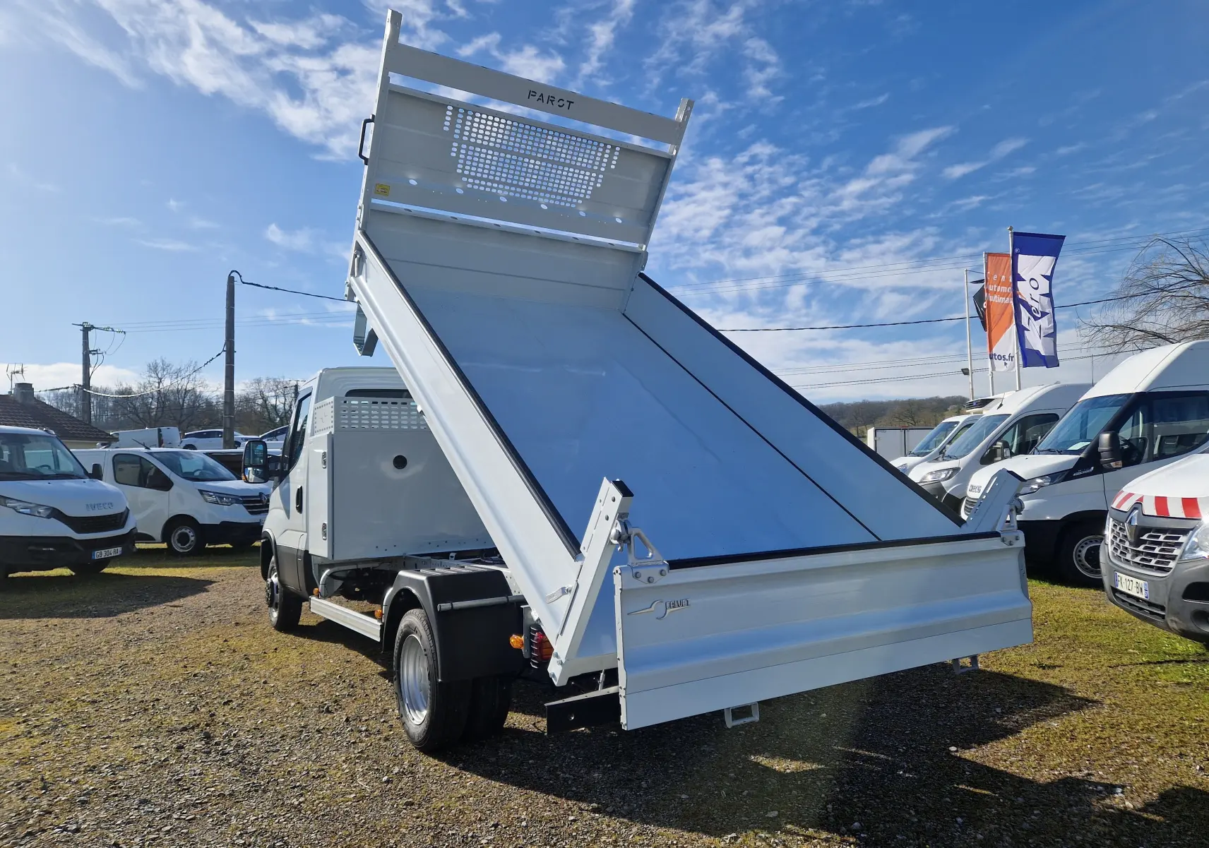 IVECO Daily blanc vu en 3/4 arrière avec benne relevée et coffre latéral visible sur terrain extérieur.