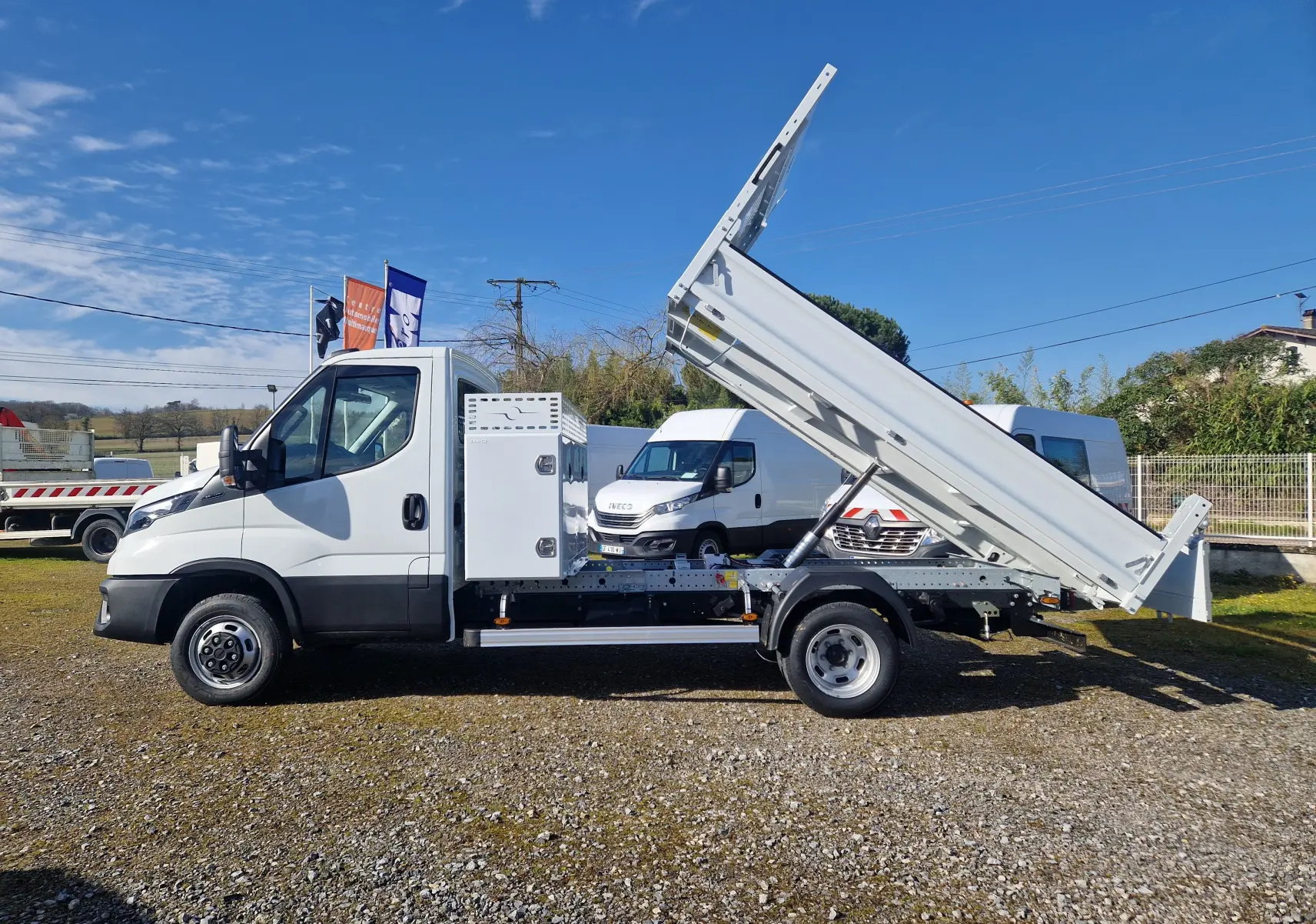 Vue latérale droite d’un Iveco Daily blanc avec benne relevée et coffre métallique fixé derrière la cabine.