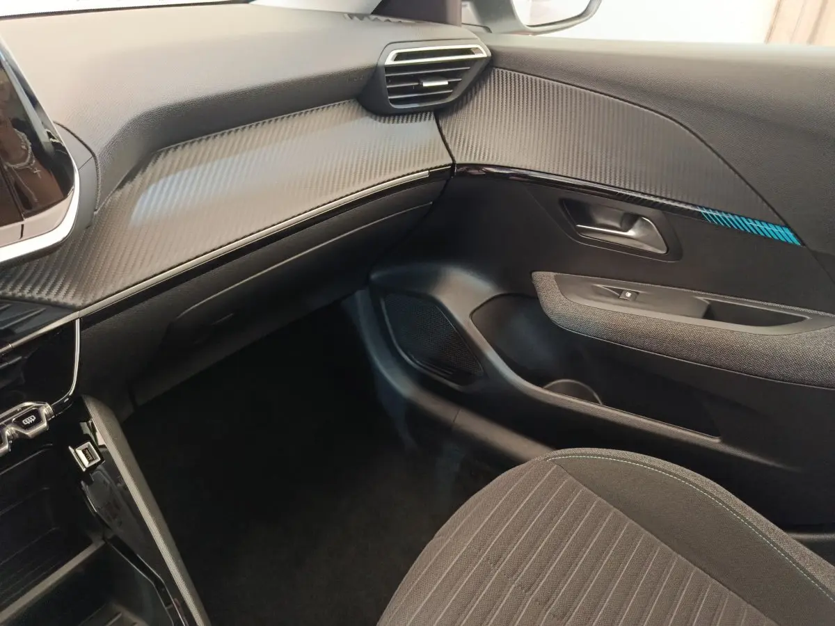 Vue intérieure côté passager de la Peugeot 208 2023, tableau de bord noir avec insert effet carbone et siège tissu gris.