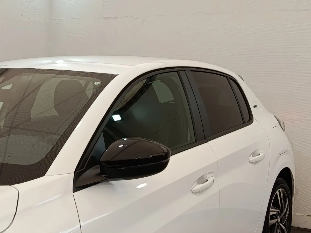 Vue de côté gauche rapprochée d’une Peugeot 208 blanche avec rétroviseur noir brillant et vitres teintées.