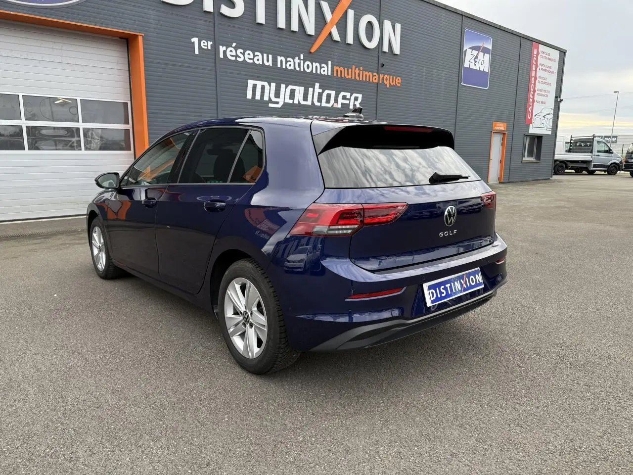 Vue 3/4 arrière droite d'une Volkswagen Golf bleu Atlantic métallisée stationnée devant un bâtiment commercial.