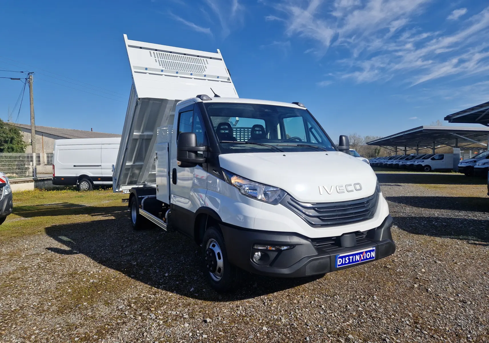 Vue 3/4 avant droit d'un Iveco Daily blanc avec benne relevée et coffre latéral visible sur un terrain extérieur.