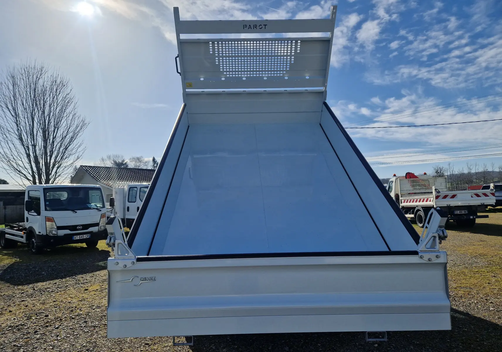 Vue arrière d’un Iveco Daily blanc avec benne acier Calvet relevée et coffre intégré, sous un ciel bleu partiellement nuageux.