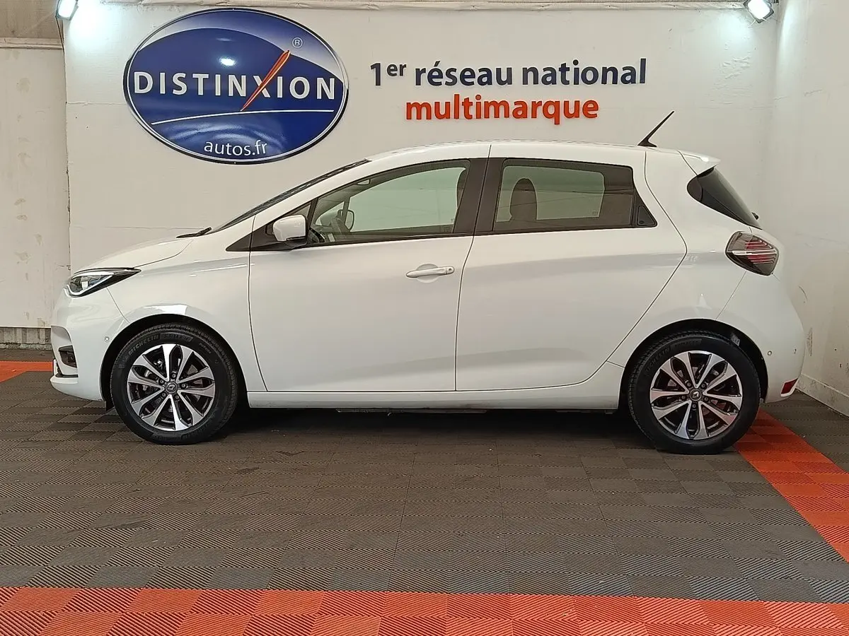 Profil droit d'une Renault ZOE Intens R110 blanche, avec jantes alliage et antenne de toit visible, en intérieur showroom.