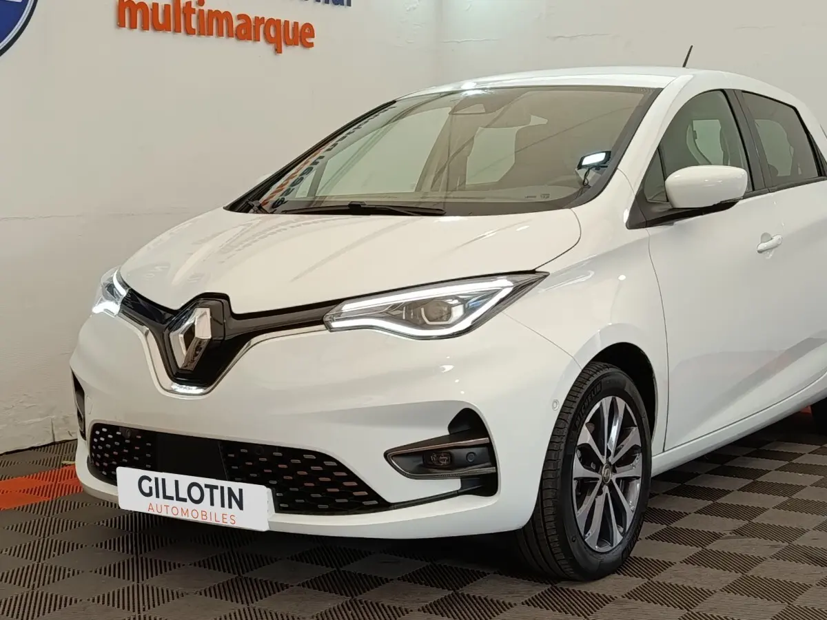 Renault ZOE Intens R110 blanc vue 3/4 avant droit, phares LED et jantes alliage visibles en intérieur showroom.