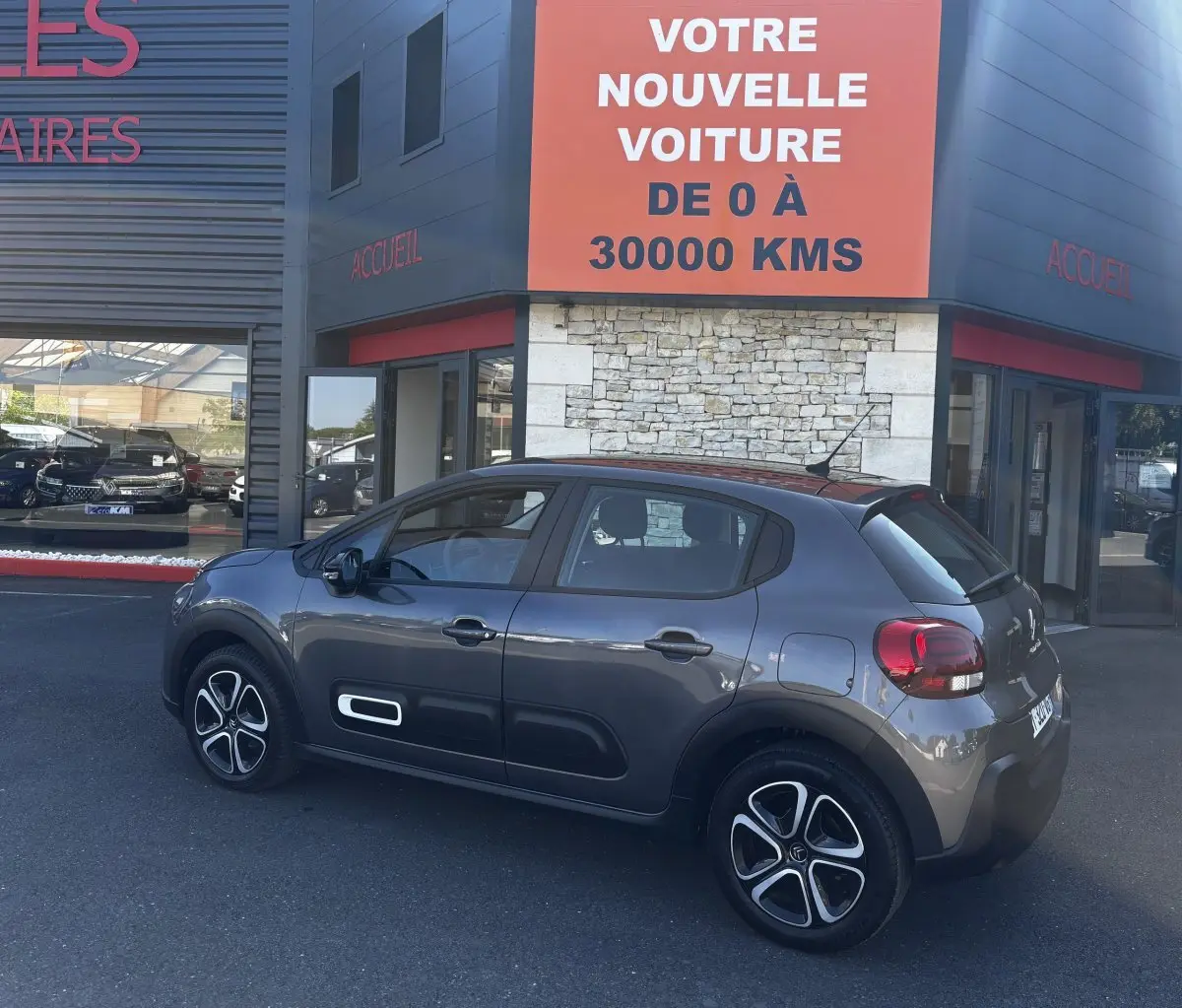 Vue côté gauche d'une Citroën C3 2024 gris foncé avec protections latérales blanches, garée devant un showroom.