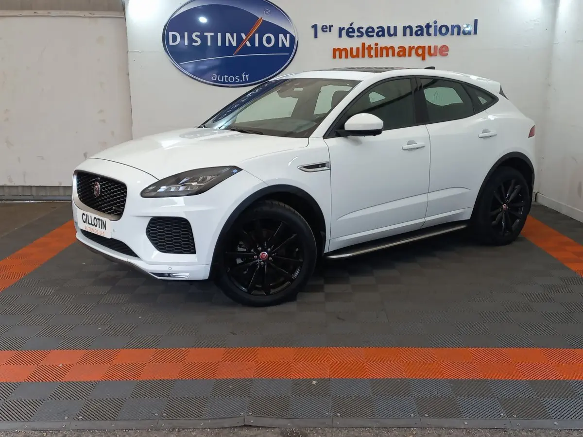 Jaguar E-PACE blanc vue 3/4 avant droit, roues noires, dans un showroom avec sol gris et bandes orange.