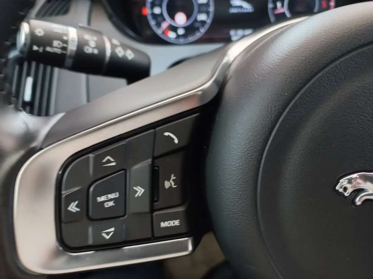 Gros plan sur les commandes gauche du volant cuir noir du Jaguar E-PACE blanc, avec logo jaguar visible.