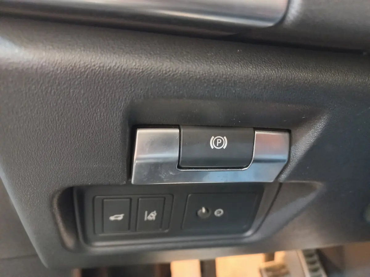 Gros plan sur le bouton de frein à main électrique et commandes du coffre dans l'habitacle noir du Jaguar E-PACE blanc.