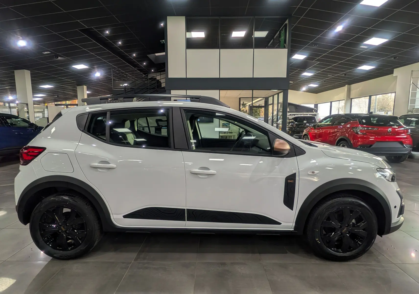 Vue latérale droite d'une Dacia Sandero Stepway blanche Glacier avec jantes noires et protections latérales noires en concession.