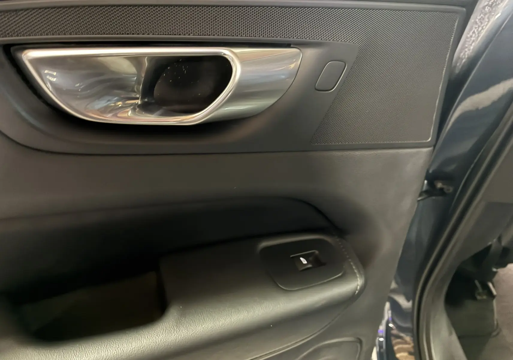 Gros plan sur la poignée intérieure chromée et la commande de vitre du côté gauche d'une porte bleu foncé du Volvo XC60 2023.