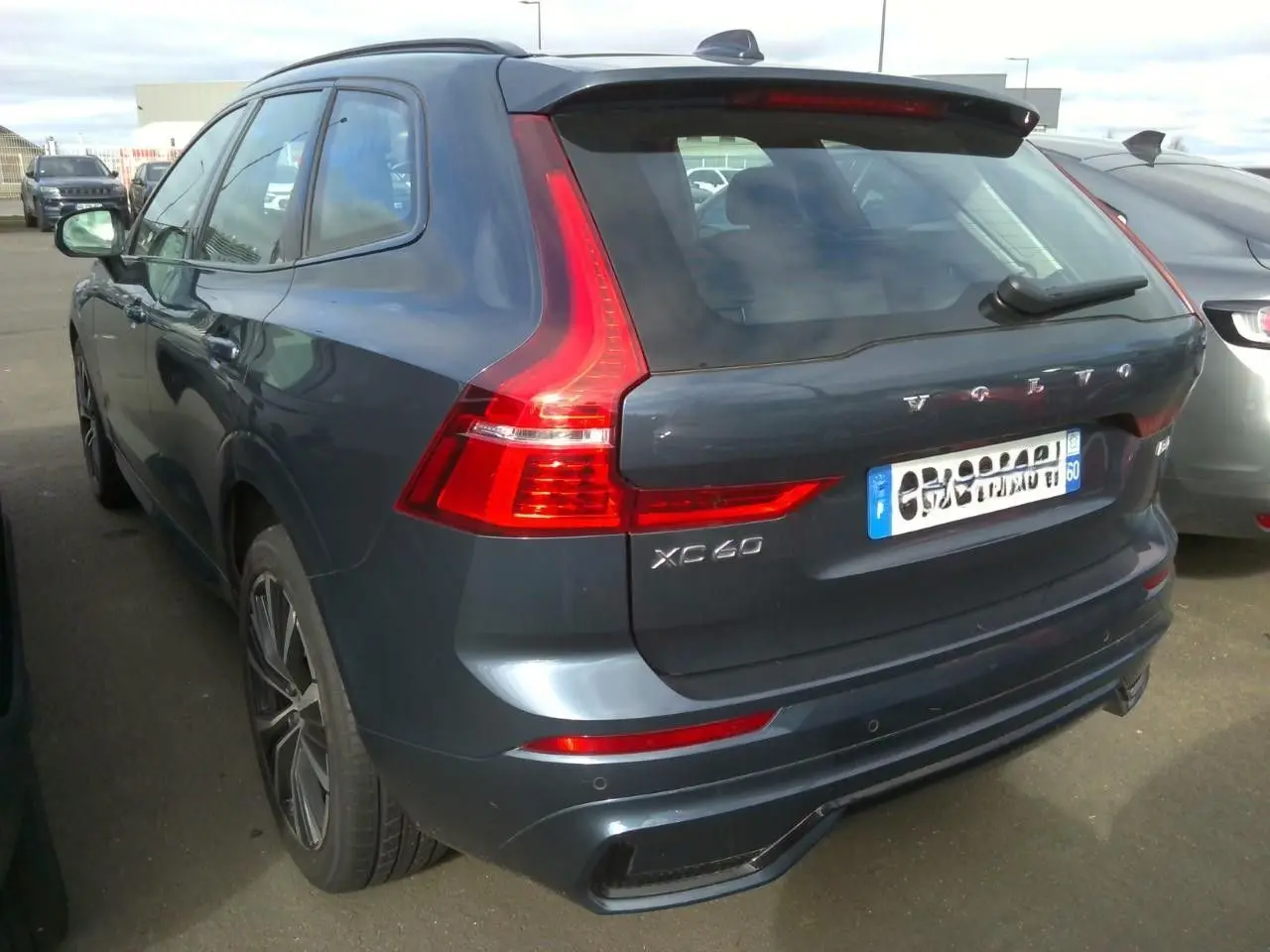 Vue frontale d’un Volvo XC60 bleu avec calandre noire laquée et phares allumés en intérieur showroom.