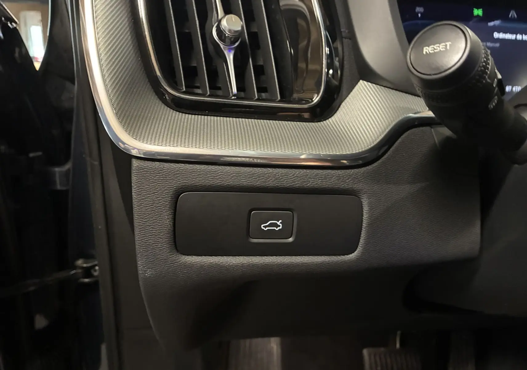 Gros plan sur le bouton d'ouverture du hayon dans l'habitacle noir du Volvo XC60 2023 côté conducteur.