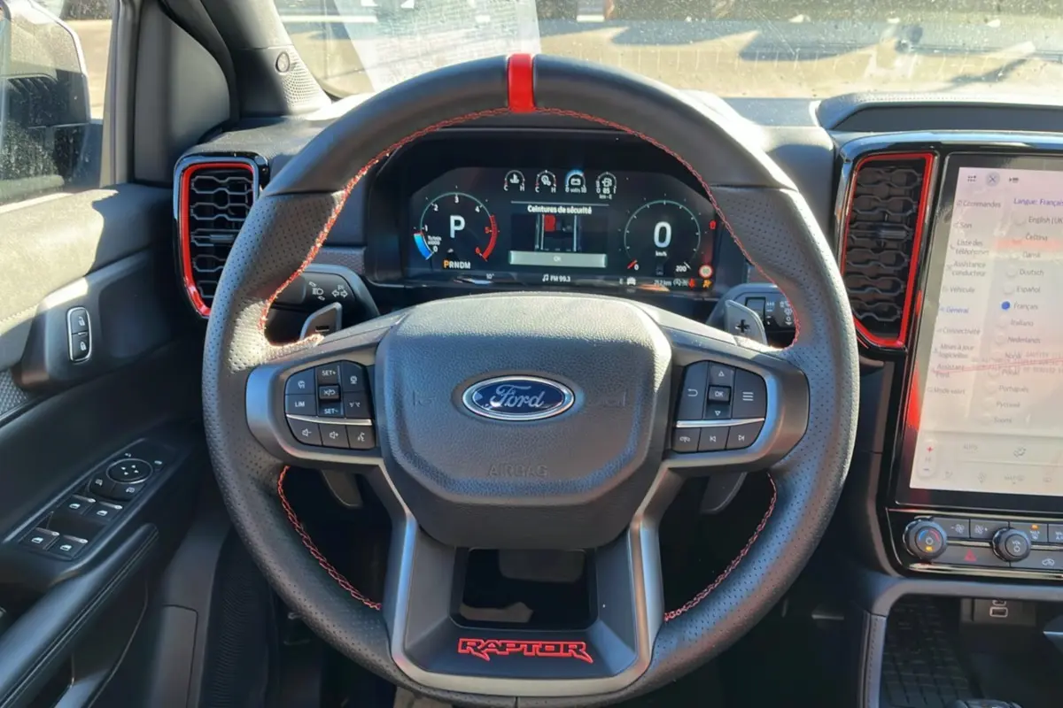 Vue intérieure centrée sur le volant cuir noir avec surpiqûres rouges et tableau de bord numérique du Ford Ranger Raptor gris commandeur.