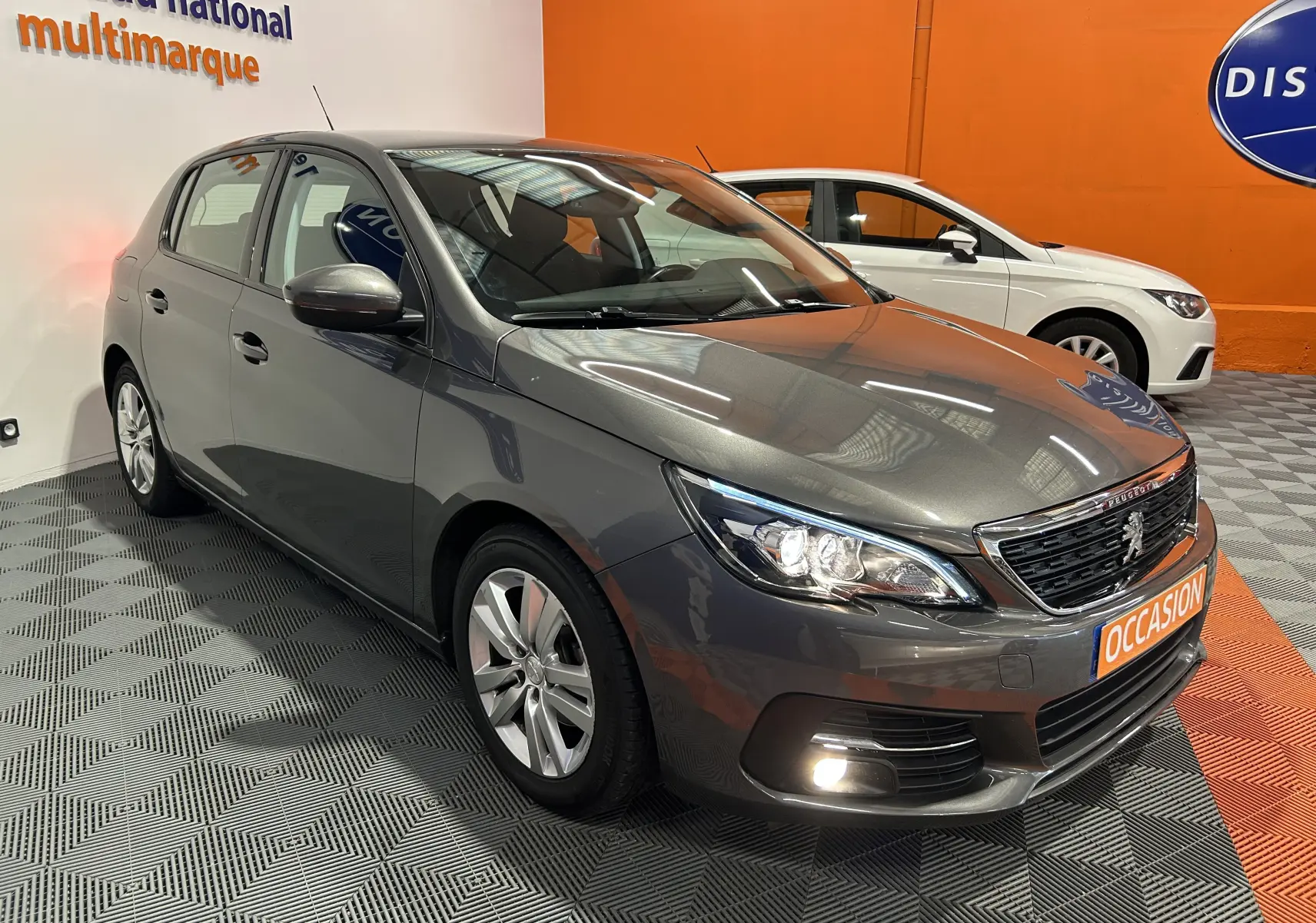 Peugeot 308 gris en 3/4 avant droit dans un showroom, avec phares allumés et plaque "OCCASION" visible.
