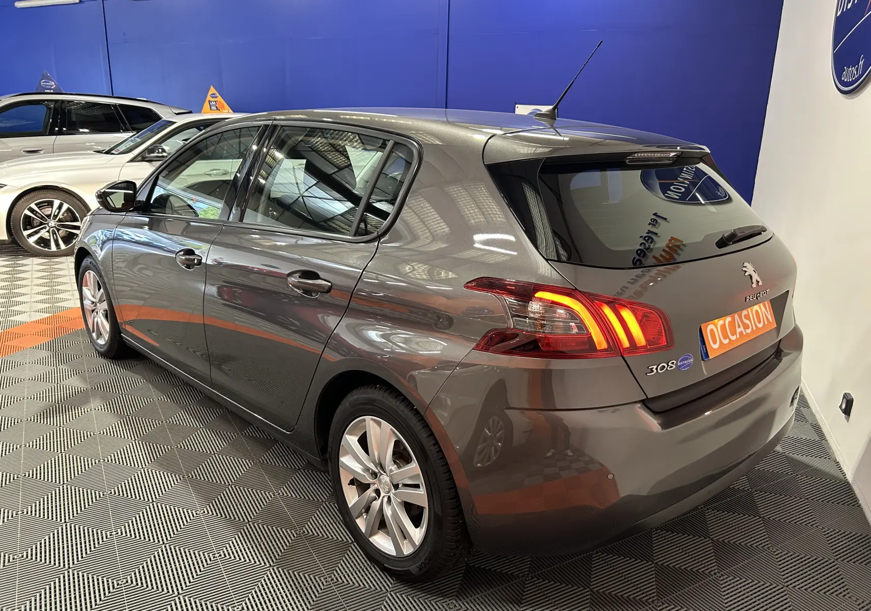 Vue 3/4 arrière droite d'une Peugeot 308 Active Pack gris métallisé, avec feux arrière allumés en concession.