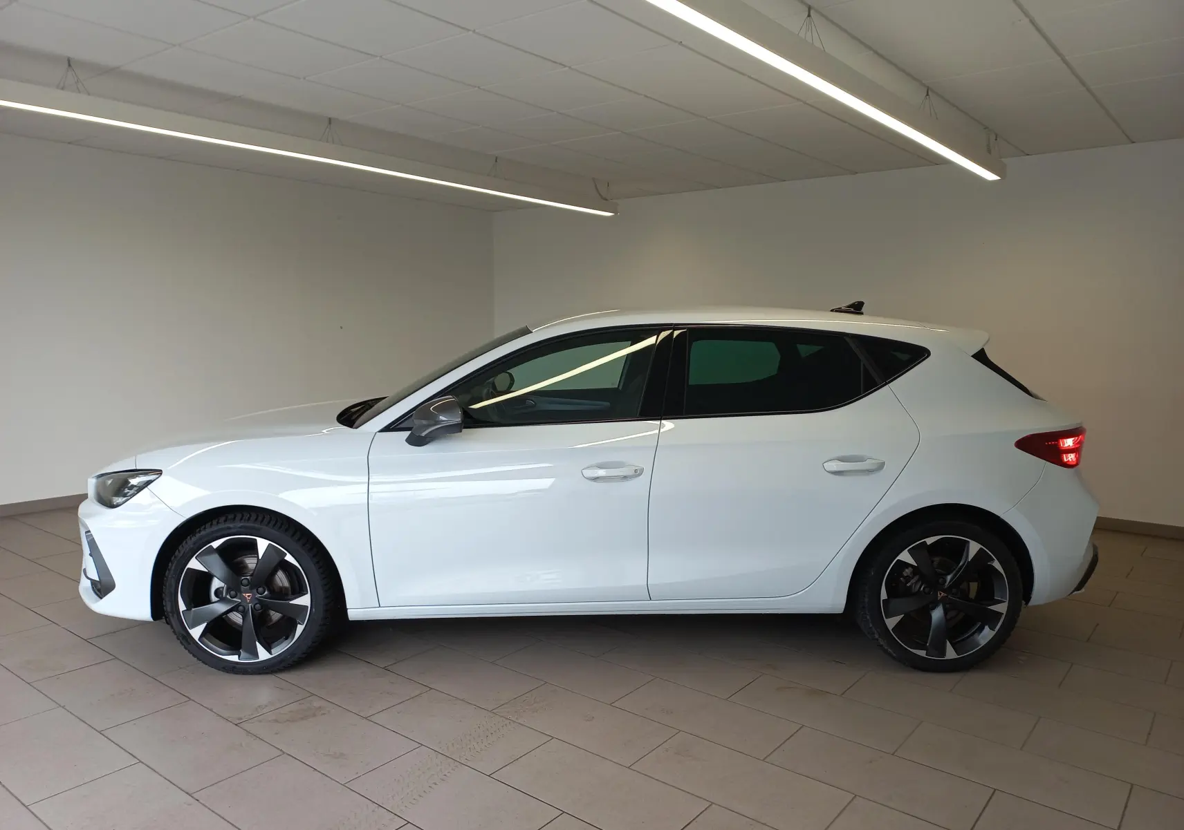 Vue de profil côté gauche d'une CUPRA Leon blanche Nevada avec jantes noires et détails cuivrés, en intérieur.