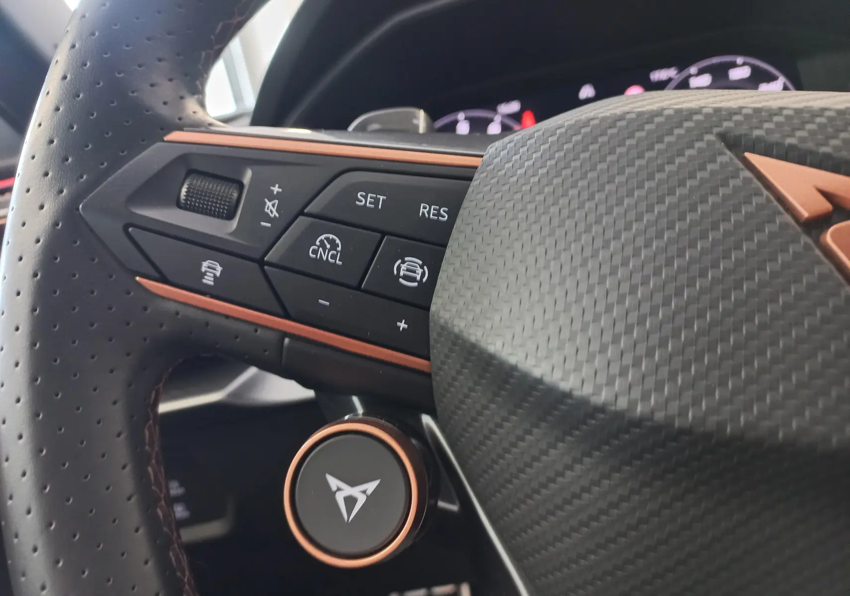 Gros plan sur la partie gauche du volant perforé en cuir avec inserts cuivrés et logo CUPRA sur fond carbone.