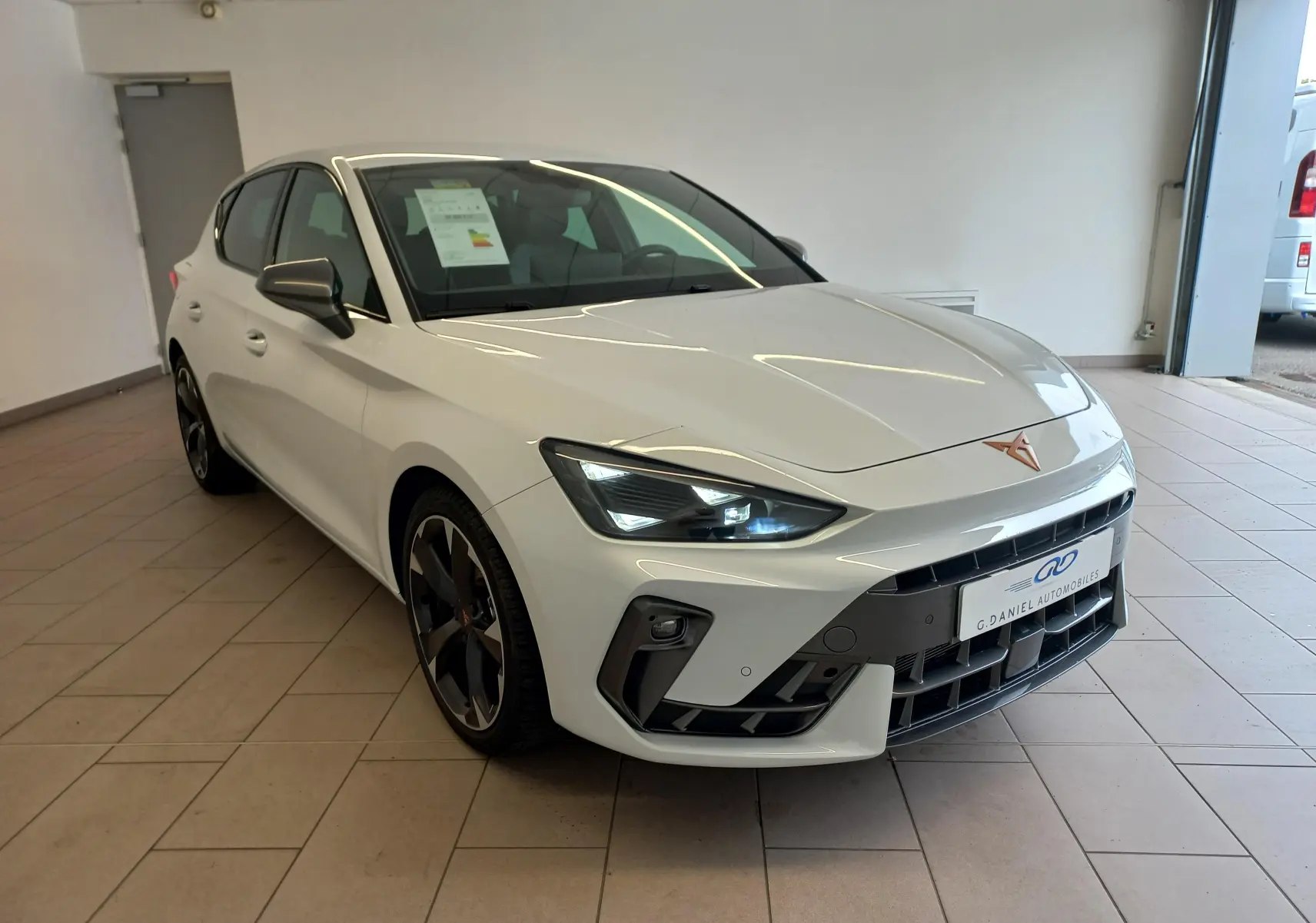 Vue 3/4 avant gauche d'une CUPRA Leon Phase 2 blanche Nevada avec jantes noires et logo cuivré sur capot.
