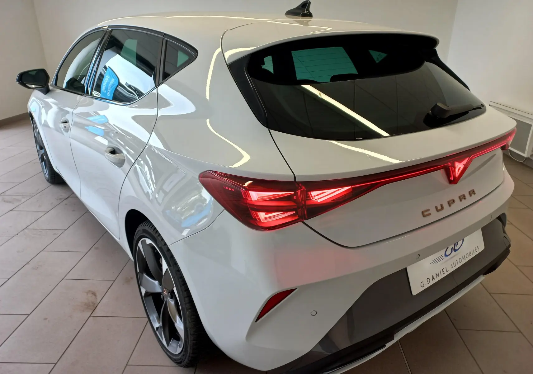 Vue 3/4 arrière droite d'une CUPRA Leon blanche Nevada avec feux arrière allumés et jantes noires en intérieur showroom.