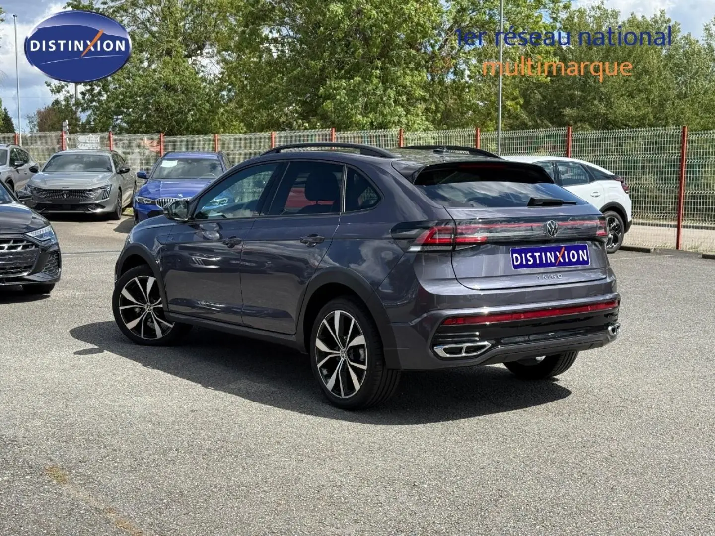 Volkswagen Taigo gris Ascot en 3/4 arrière droit, avec jantes alu et barres de toit visibles sur parking.