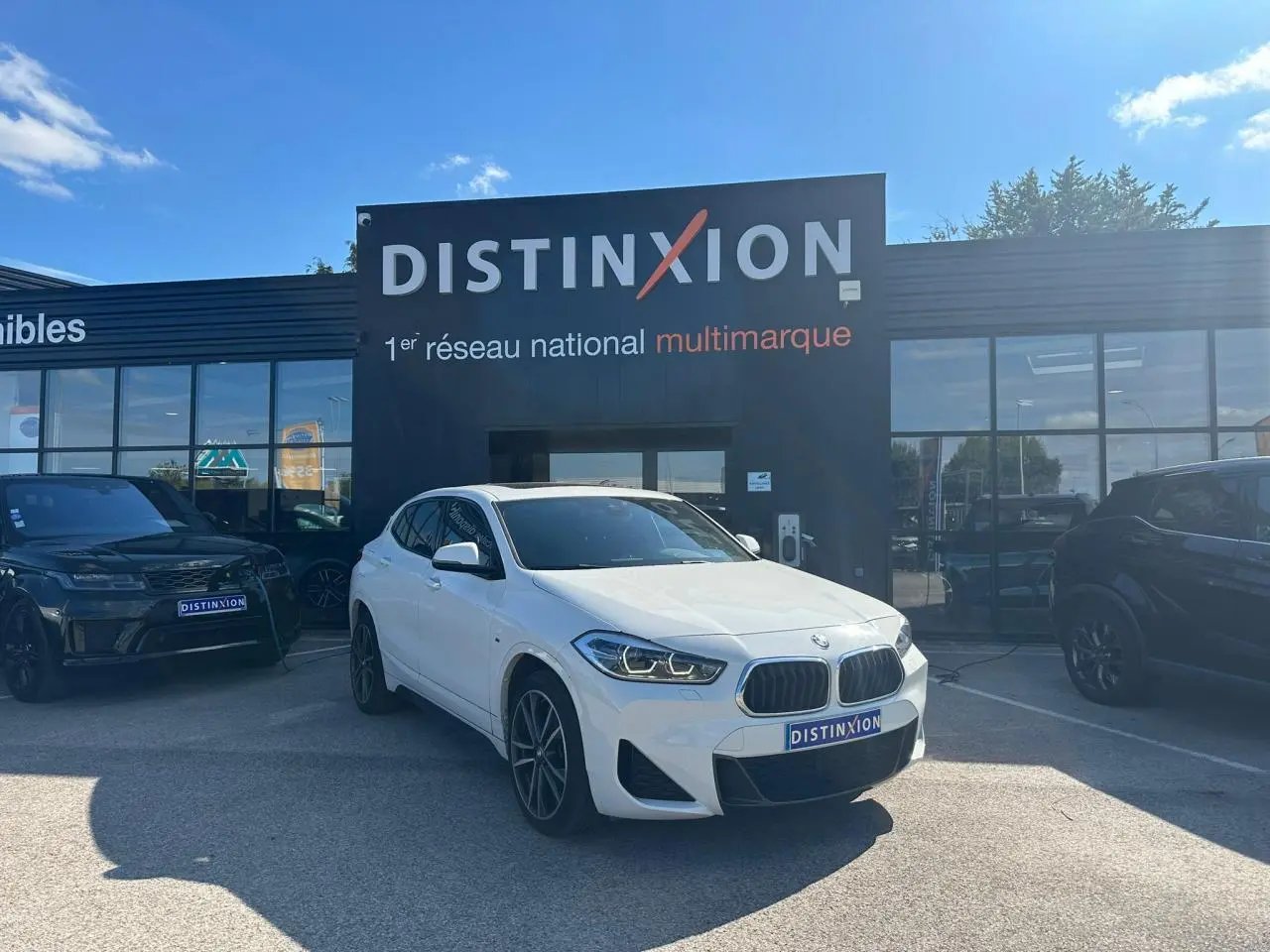 BMW X2 blanc alpin vue 3/4 avant droit avec jantes M 19 pouces devant un showroom Distinxion