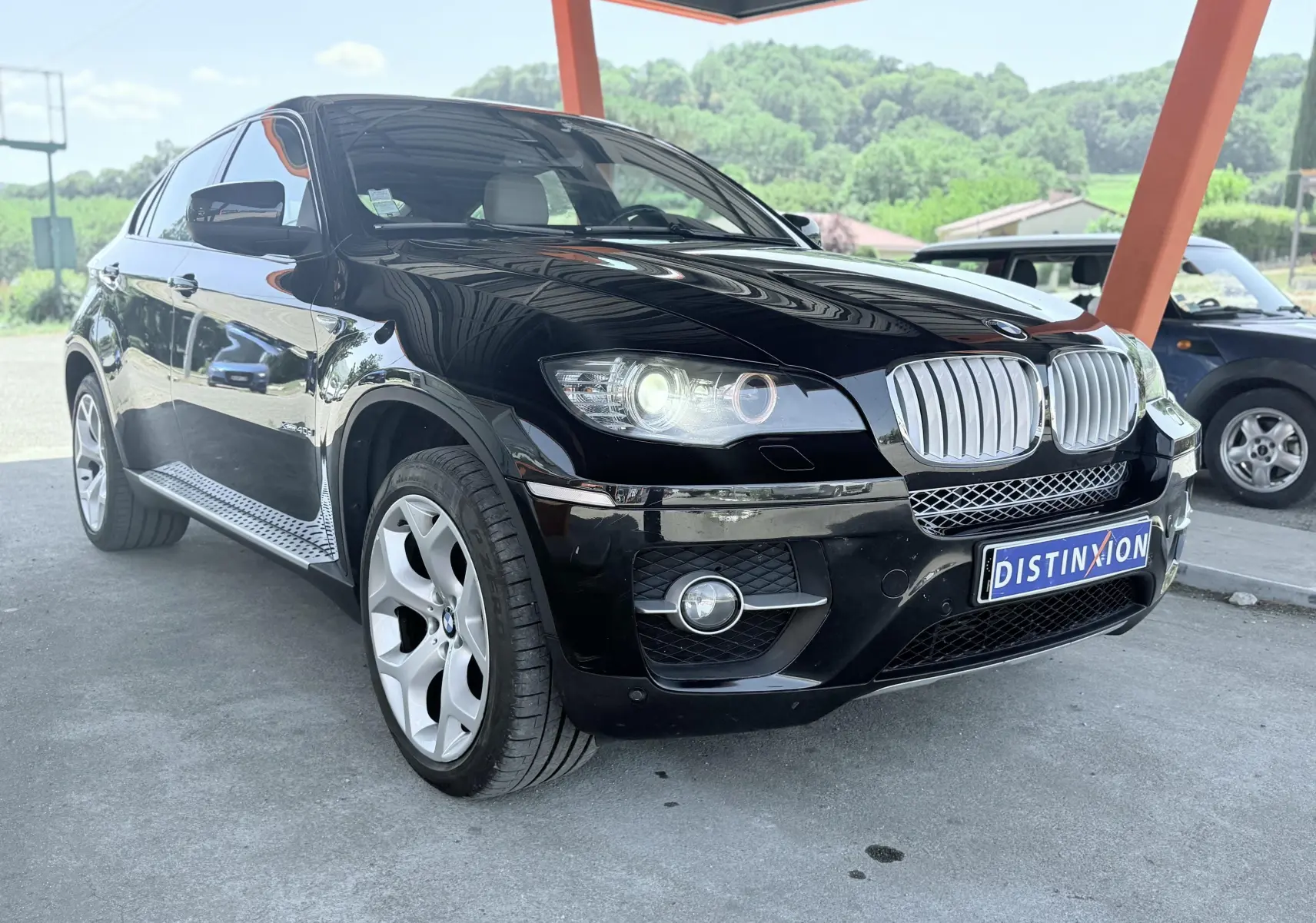 BMW X6 noire vue en 3/4 avant droit, avec calandre chromée et phares allumés sous un abri orange.