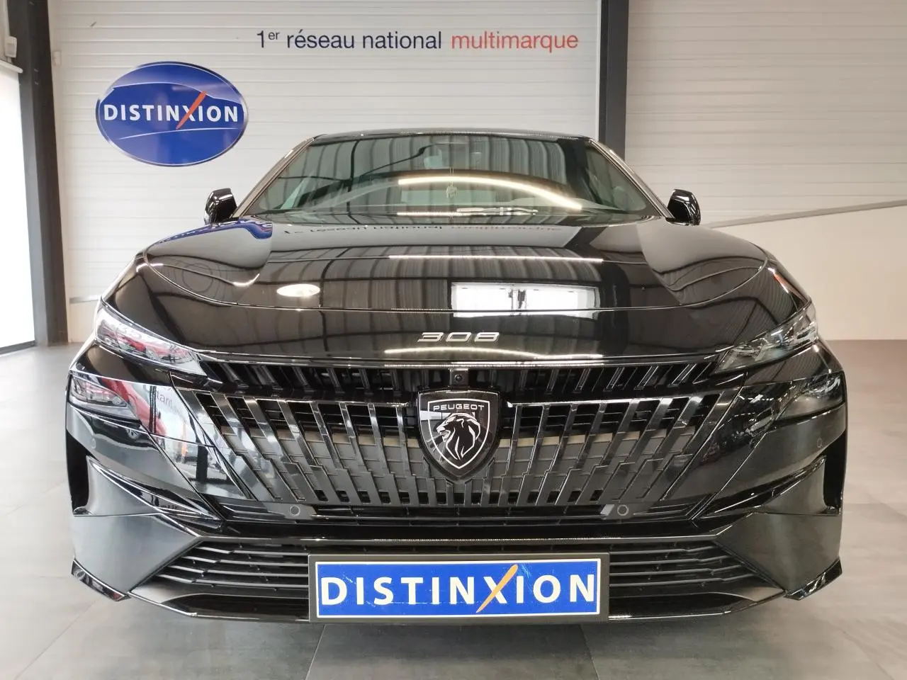 Vue frontale d'une Peugeot 308 noire métallisée avec calandre et blason Peugeot distinctifs, en intérieur showroom.