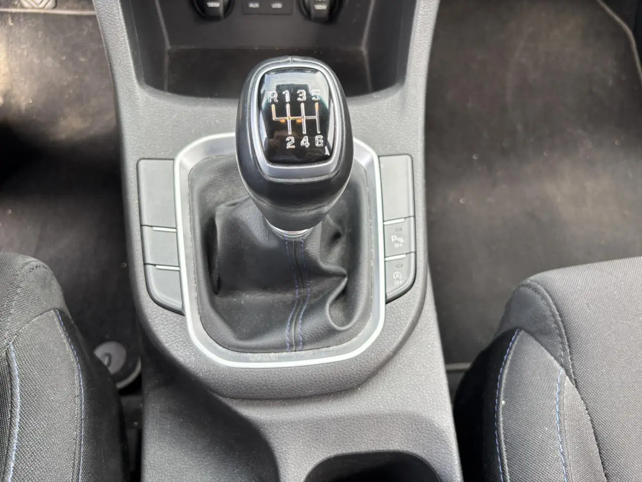 Gros plan sur le levier de vitesses manuel noir avec surpiqûres bleues, entre les sièges avant noirs de la Hyundai i30 blanche.