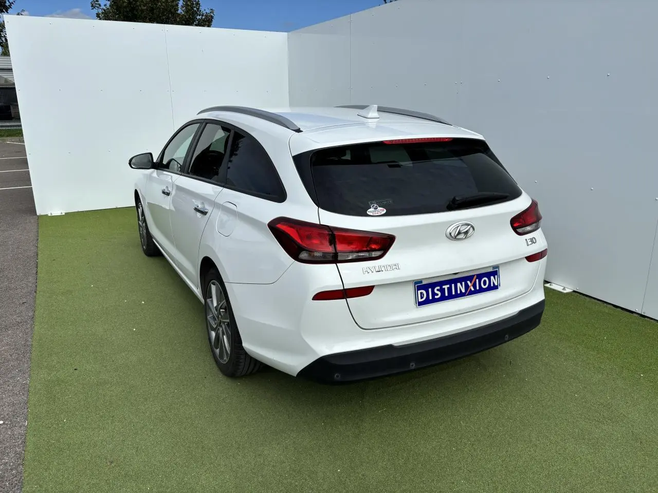 Vue 3/4 arrière droite d'une Hyundai i30 break blanche avec vitres arrière surteintées et barres de toit noires