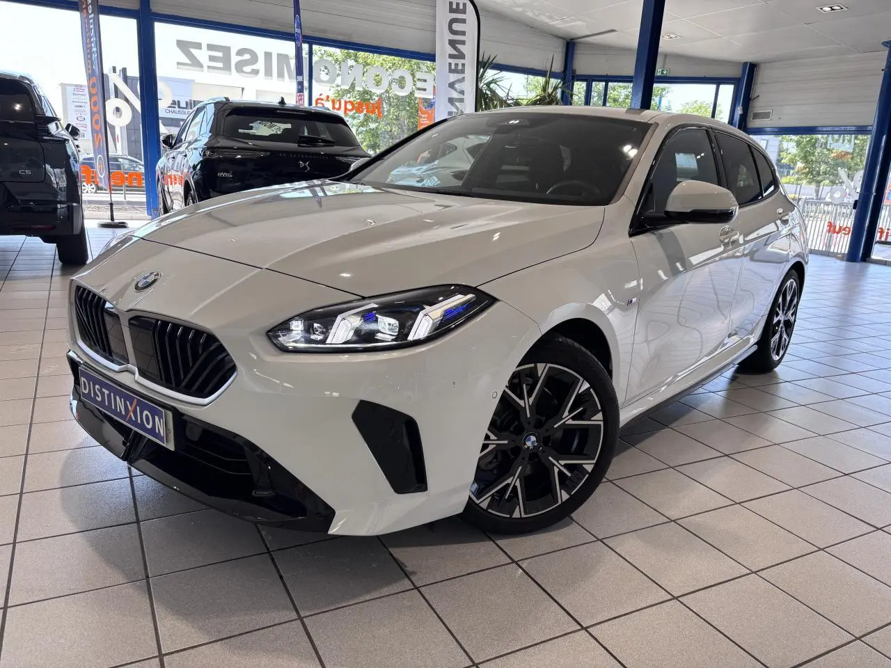 BMW Série 1 120d M Sport blanche vue 3/4 avant droit en showroom, avec jantes noires et calandre noire distinctive.
