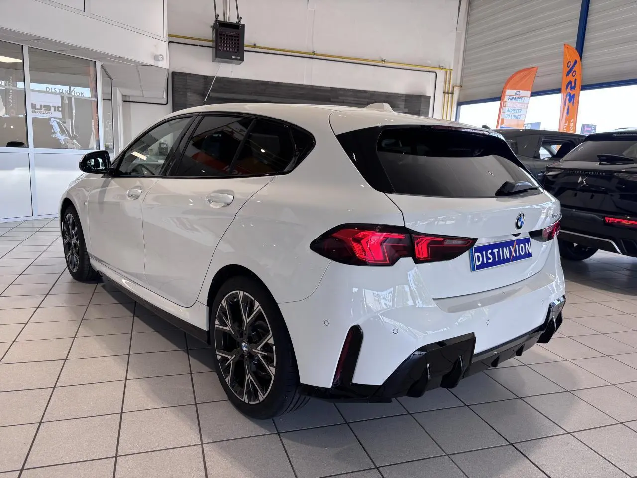 Vue 3/4 arrière droite d'une BMW Série 1 120d M Sport blanche avec jantes noires en concession.