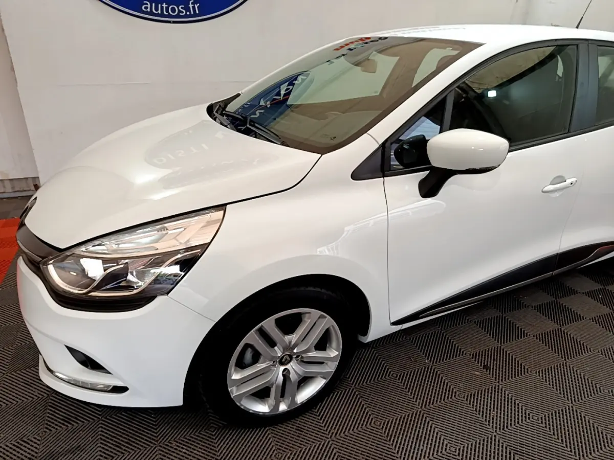 Renault Clio Business TCe 90 blanc vue 3/4 avant droit, phares LED et jantes alliage visibles sur fond intérieur.