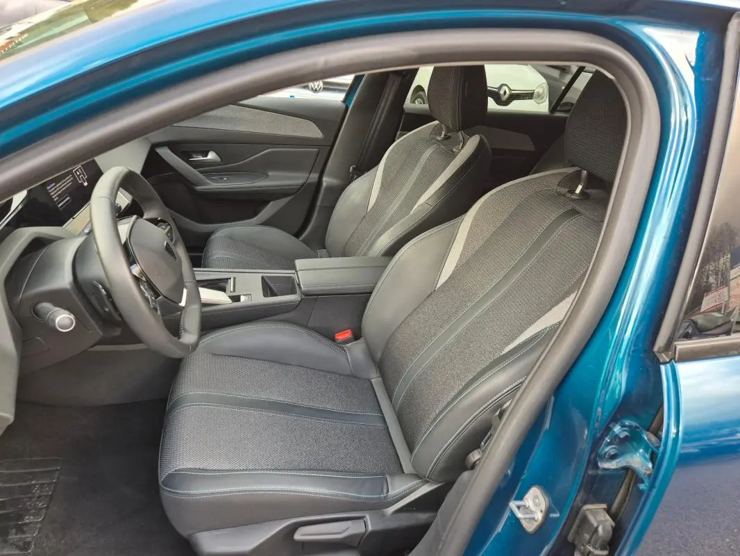 Vue intérieure côté conducteur d'une Peugeot 308 bleu 2024, sièges tissu et cuir avec accoudoir central et cockpit numérique.